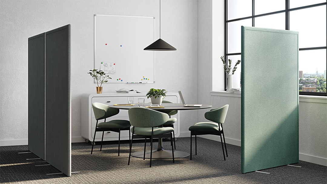 Light mint green acoustic office divider screen, non-linking, 42mm thick covered on both sides in sound absorbing fabric, with free-standing feet, separating a bright room in two spaces, next to a round table with three meeting chairs and a large window